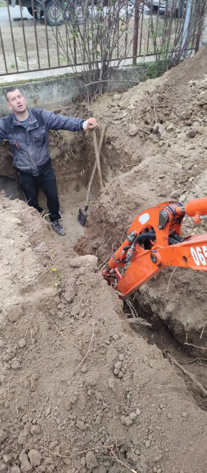 Radnik u iskopanom rovu pored bagera koji kopa kanal uz ogradu
