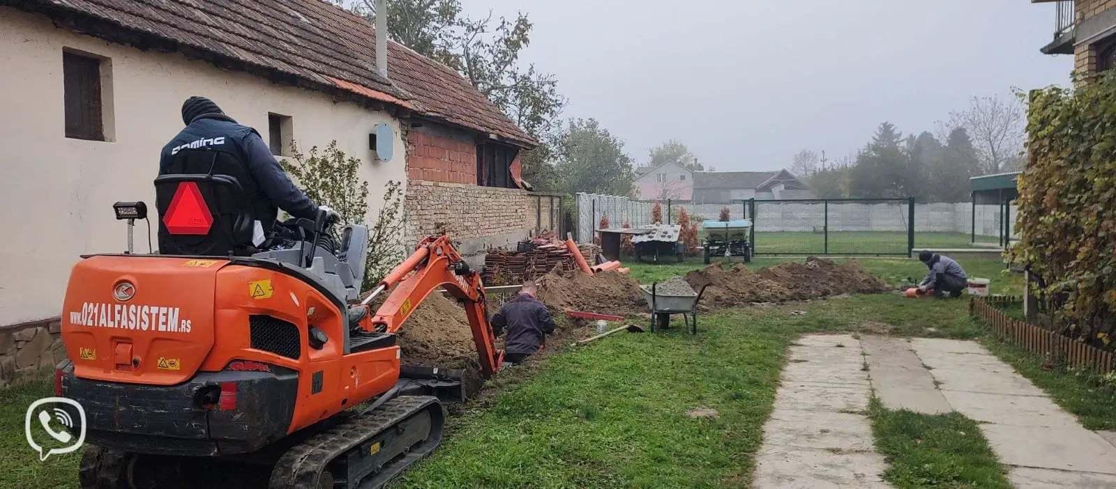 Mini bager 021 Alfa Sistem i radnici na iskopu kanala duž stare kuće